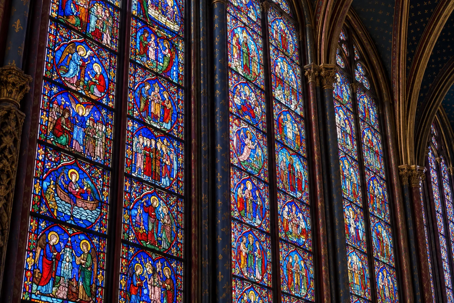 Détail de vitraux médiévaux où les scènes figurées se dessinent dans des panneaux rouges et bleus.