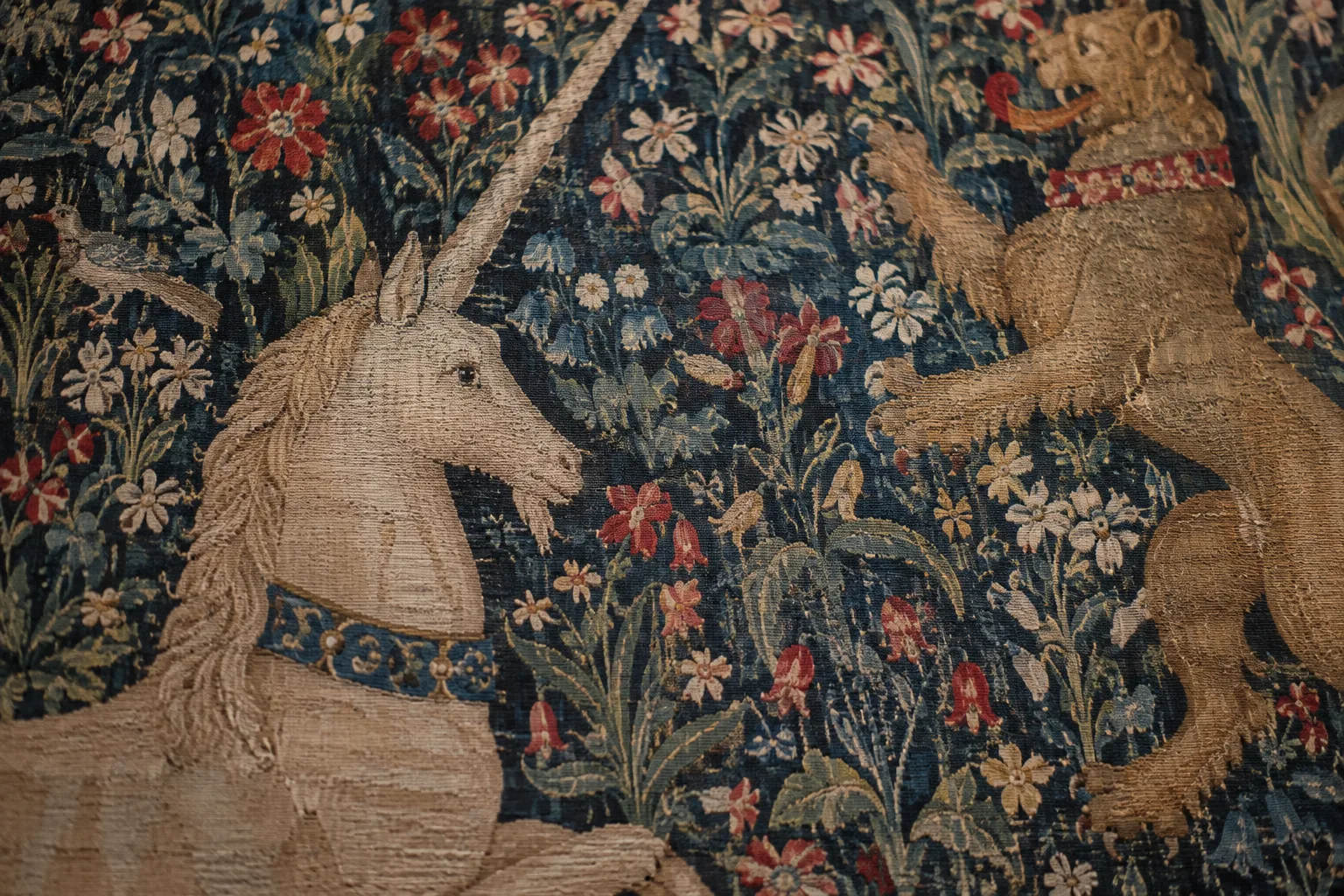 Détail d’une tapisserie médiévale aux motifs de fleurs et d’animaux, éclairée dans une salle de musée.